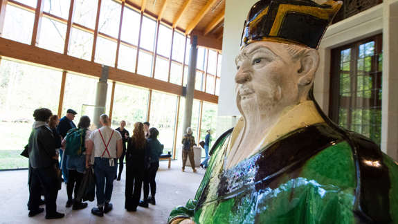 Large ceramic statue of a Chinese official in green and yellow robes in the foreground, with a group of visitors gathered in the background inside a gallery with tall windows and wooden beams.