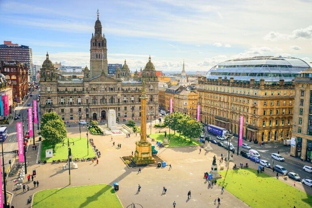 A birds eye view of George Square. It's a sunny day, and people are walking around the square. Glasgow City Chambers and Sir Walter Scott Monument can be seen.