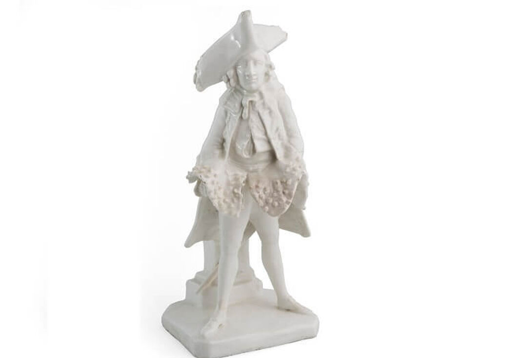 A white porcelain statue of a man dressed in 18th Century fancy garb that includes a flamboyant hat.