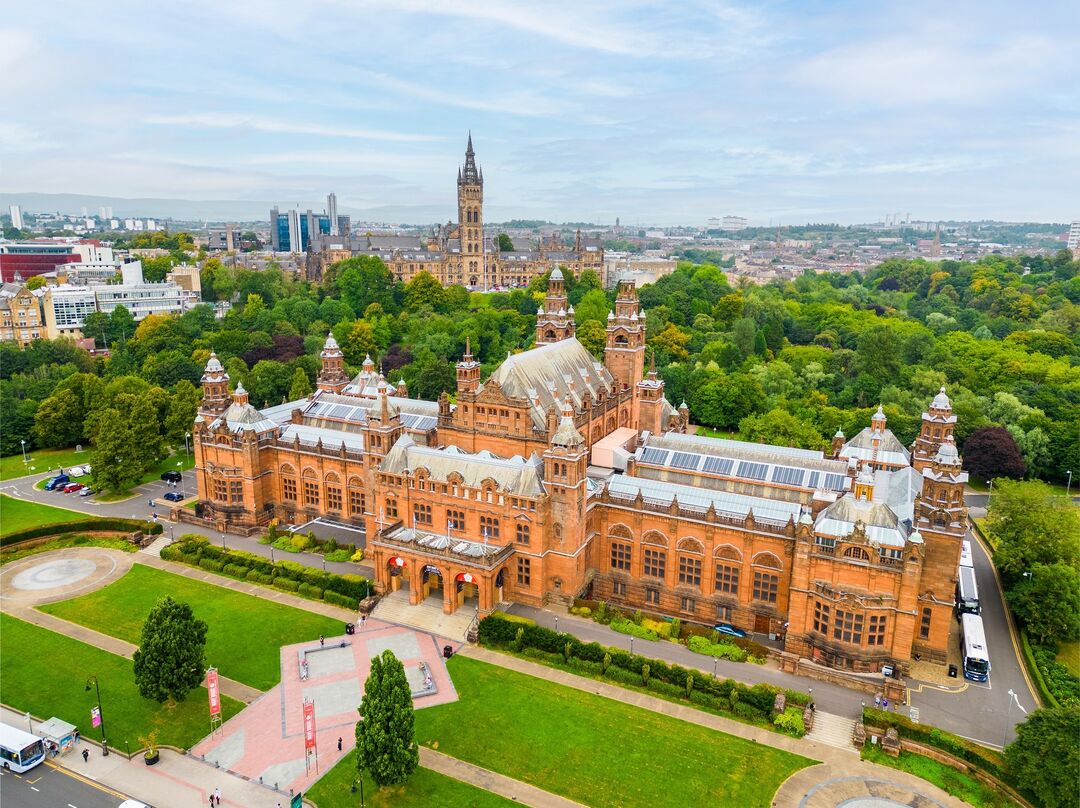 Drone view of a grand red building surrounded in greenspace, with a grand building with a spire and hills in the distance.