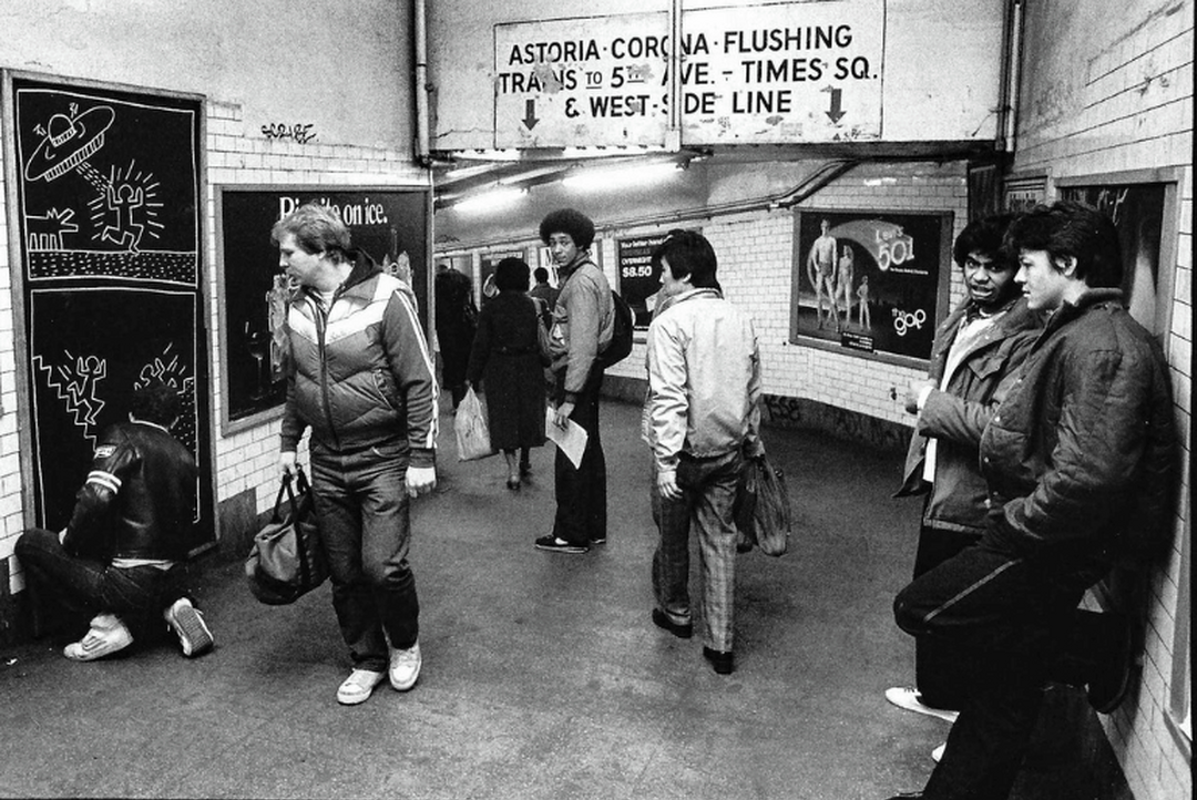 A black and white image of a group of people in a subway going about their lives. One person is drawing on a board and people are interested in what they're doing.