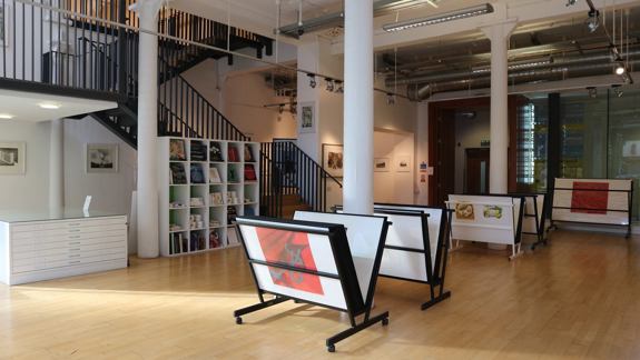 An empty art studio and gallery with a light wooden floor. The space has a staircase, white support columns, and racks displaying framed artwork.