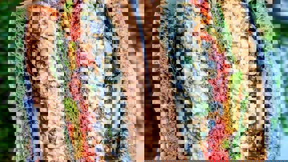 A close-up of a thick sandwich, sliced in half, revealing a creamy egg salad filling with avocado, tomato, and lettuce, on wholemeal bread.