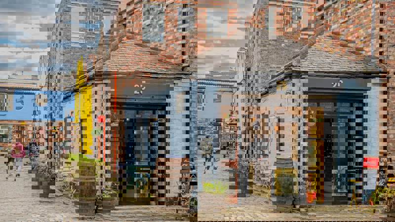 A brick and light-blue painted cafe with a small outdoor seating area sits in a cobbled alleyway.