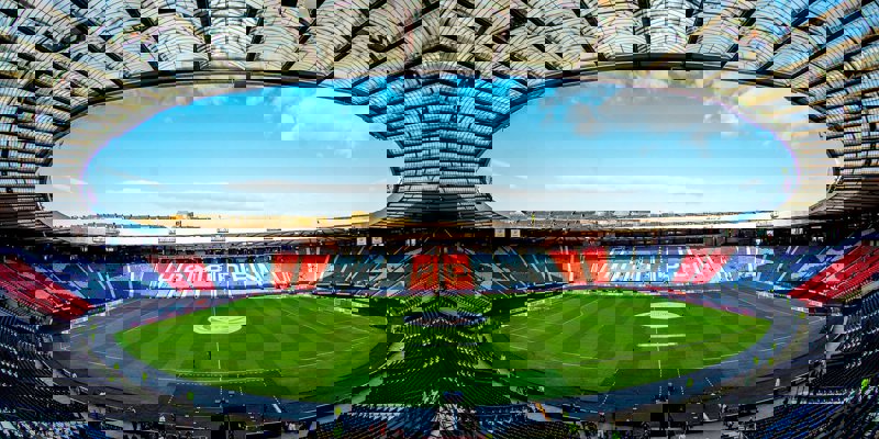 An empty Hampden Park stadium with blue and red seats, a green field, and a transparent roof against a blue sky.