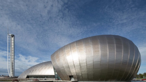 The exterior of the Glasgow Science Centre, with its two curved, silver buildings and tall tower, stands next to water.