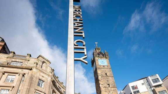 A modern, tall sign with the words 'Merchant City' on it, standing in front of the historic Tolbooth Clock Tower in Glasgow.