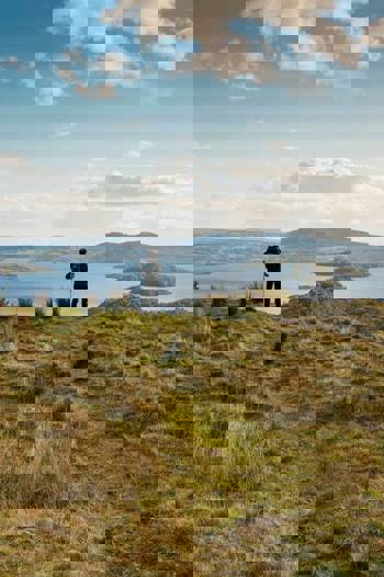 A hiker stands on a grassy hill overlooking a vast loch dotted with islands, with rolling hills in the background