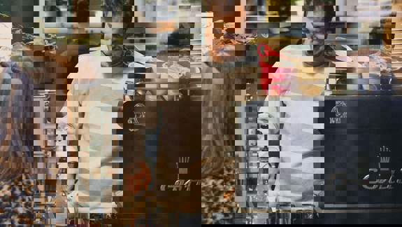 A barista in a striped sweater smiles while preparing drinks at a coffee machine, with a customer visible.