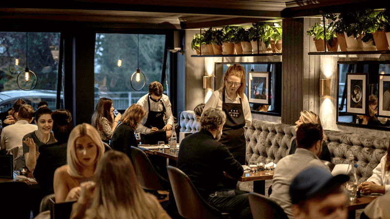 Busy restaurant interior at dusk, with diners, waitstaff, upholstered seating, potted plants, framed wall art, and large windows providing natural light.
