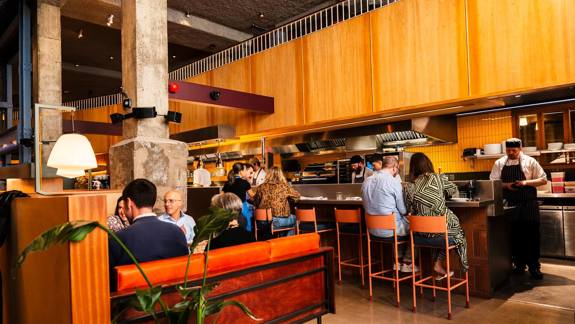 The interior of a modern restaurant with a counter, people seated at tables and a chef working in the open kitchen.