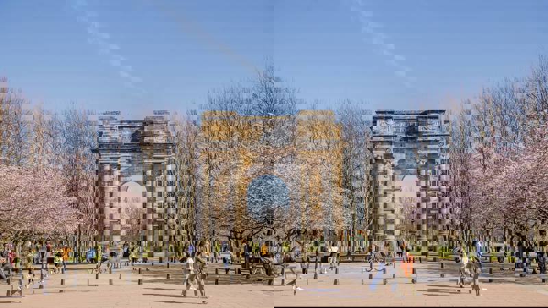 People walk near the grand McLennan Arch in Glasgow Green, surrounded by cherry blossom trees in full bloom on a sunny day.