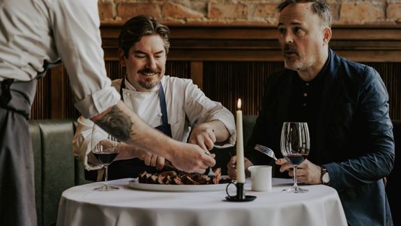 A chef and a diner sit at a candlelit restaurant table as a server slices and serves a large steak; exposed brick walls and glasses of red wine create a warm bistro atmosphere.