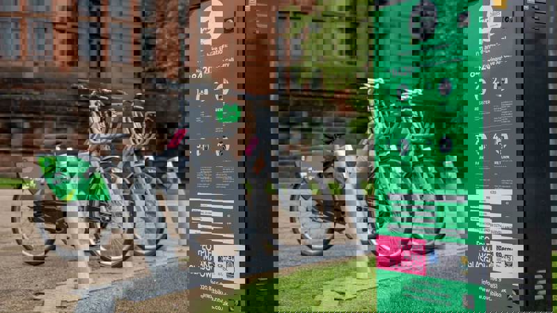 Two Ovo e-bikes are parked at a hire station in front of the Kelvingrove Art Gallery.