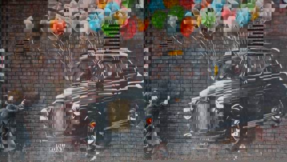 A street mural of a black taxi cab being lifted by colorful balloons, with a person on the left seemingly letting go of the balloons.