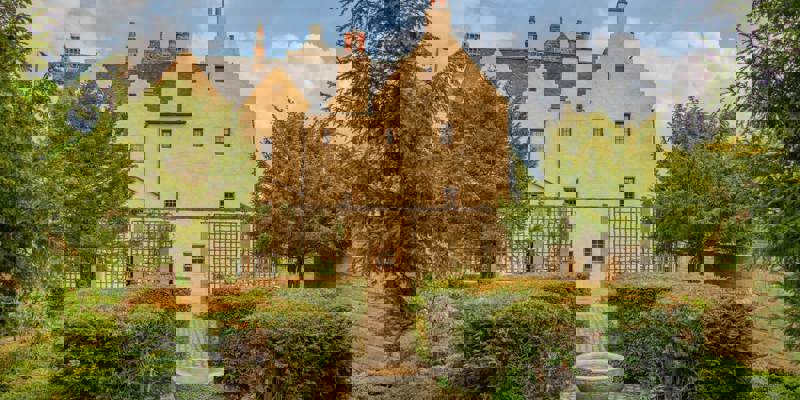 A historic, light-colored building with multiple gables and windows, surrounded by a manicured garden with a stone birdbath.