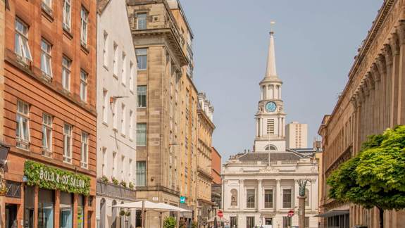 A wide street view showing historic architecture in Glasgow, including a church with a tall steeple and clock face, and several cafes.