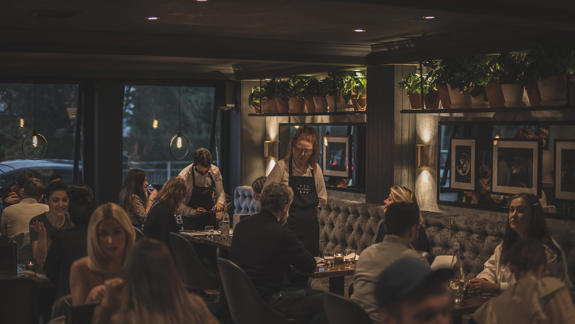 A dimly lit, stylish restaurant interior with patrons seated at tables and a server wearing a black apron.