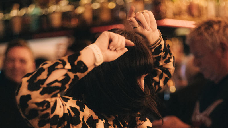 A person with dark hair in a leopard print shirt is dancing with their arms raised in a dimly lit bar.