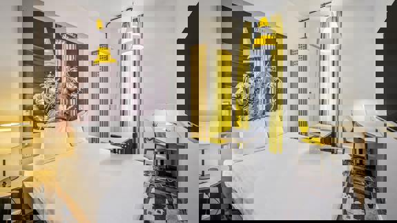 Bright modern bedroom at ibis Styles Glasgow Centre George Square featuring a large bed, yellow accents, and wall art of the Duke of Wellington statue with a traffic cone.