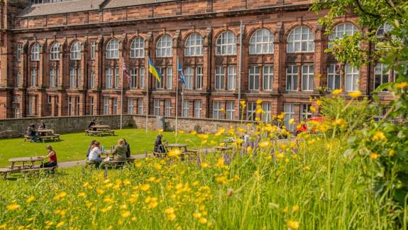 Students sit on picnic benches in a garden filled with yellow wildflowers, in front of a large red brick building.