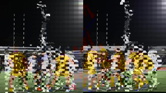 An exciting night-time rugby lineout, with a player leaping high to catch the ball above a cluster of players in yellow and dark jerseys.