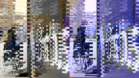 A person in a wheelchair is using the ramp to board a bus with "First Bus" written in large white letters on its purple side.