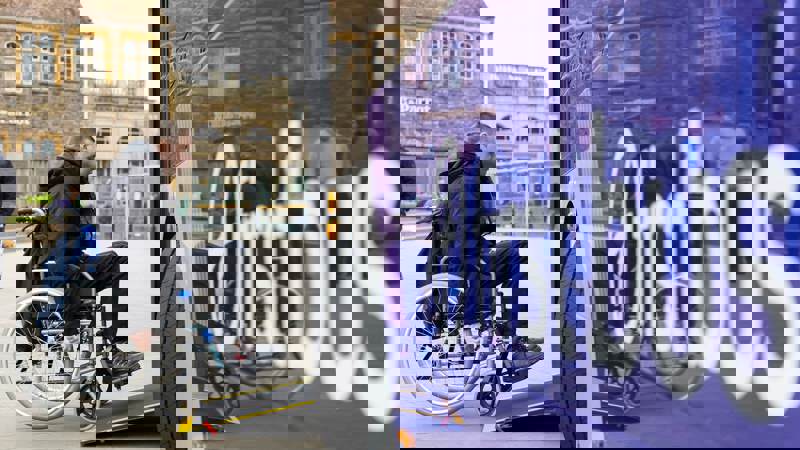 A person in a wheelchair is using the ramp to board a bus with "First Bus" written in large white letters on its purple side.