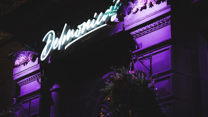 An exterior shot of a building at night, with a bright purple neon sign that reads 'Delmonica's' shining above the entrance.