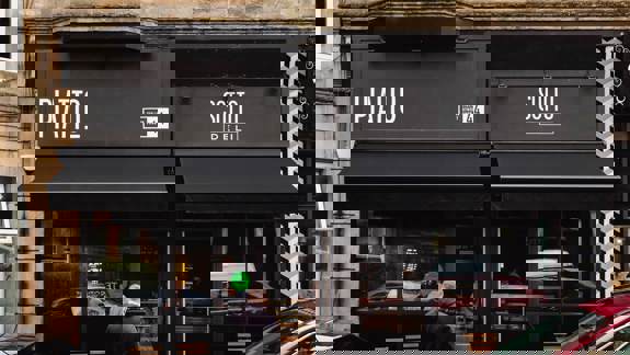 A dark storefront with awnings for two businesses, Piatto and Sotto Deli. A man in a dark jumper stands outside, and several people are visible inside. A red car is parked on the street in front of the building.