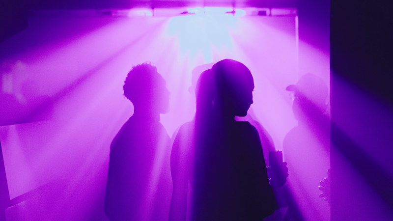 Backlit silhouettes of people standing together in a room illuminated by vibrant purple light beams creating a dramatic, atmospheric scene.