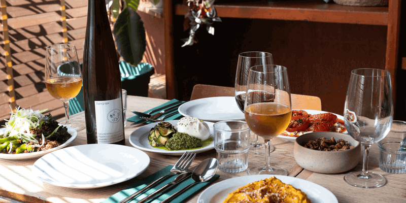 Dining table with salad, courgette and cheese dish, roasted tomatoes, mashed dish with herbs, wine glasses, water, wine bottle, green napkins, and plants in background.
