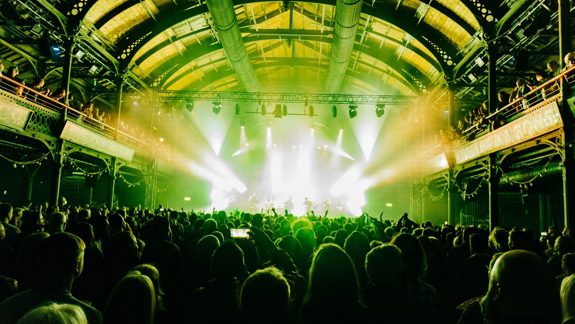A crowded music venue with a band performing on a stage under bright spotlights, with the audience silhouetted in the foreground.