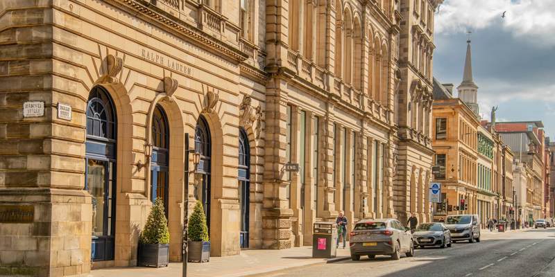 A sunlit street scene in Glasgow with a large, ornate building housing a Ralph Lauren store and traffic moving along the road.