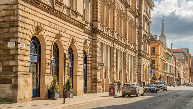 A sunlit street scene in Glasgow with a large, ornate building housing a Ralph Lauren store and traffic moving along the road.