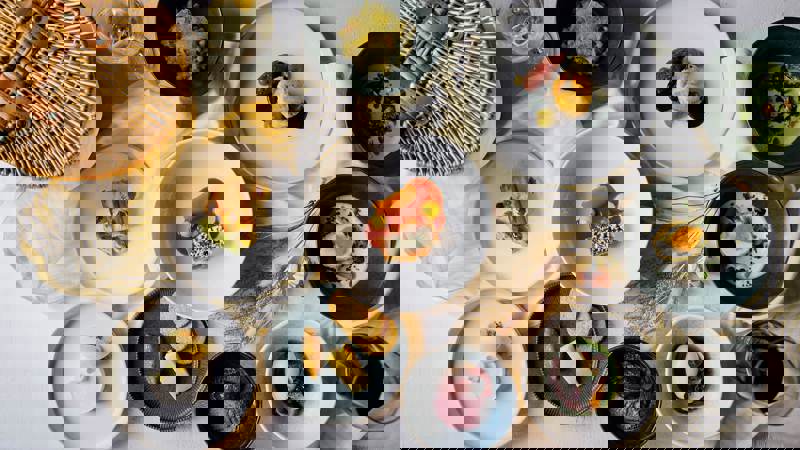 A variety of gourmet dishes are displayed on a white tablecloth, including a cheeseboard, appetizers, and several small plates. A picnic basket and glasses of wine are also visible.