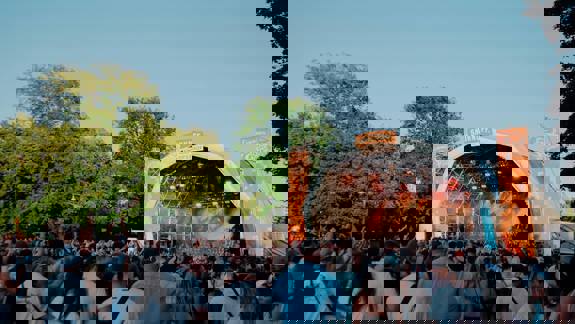 A wide-angle shot of a music festival stage during the daytime, with a crowd of people watching the performance.