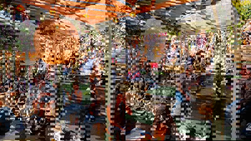 Outdoor seating area with tiered wooden benches under a clear roof, filled with people enjoying drinks