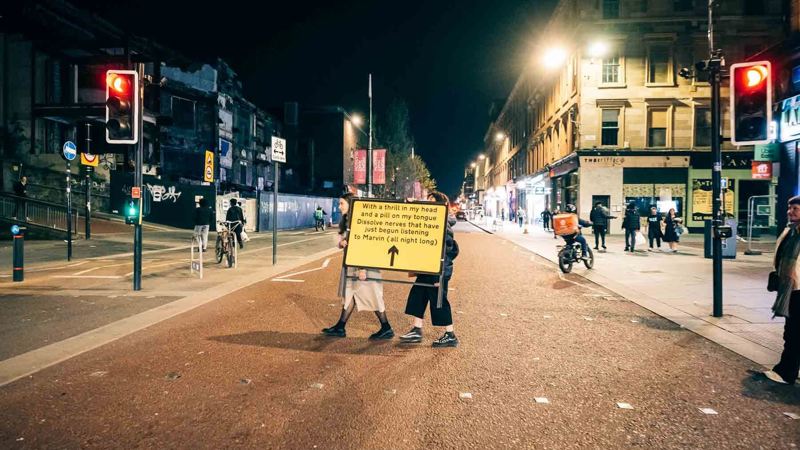 People walk at night across Sauchiehall Street holding a large yellow sign with text. Illuminated buildings and traffic lights create an urban ambience.