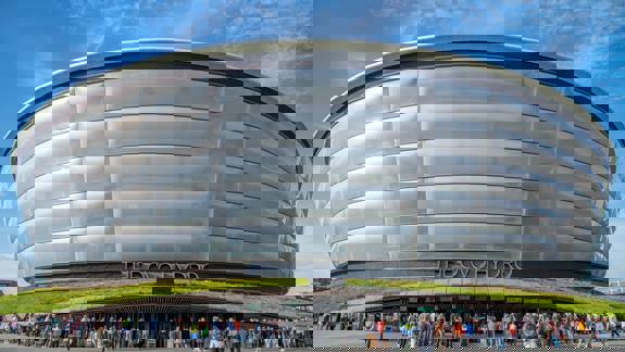 A large crowd of people gathers in front of the OVO Hydro, a large, modern-looking, circular arena with a ridged, translucent exterior.