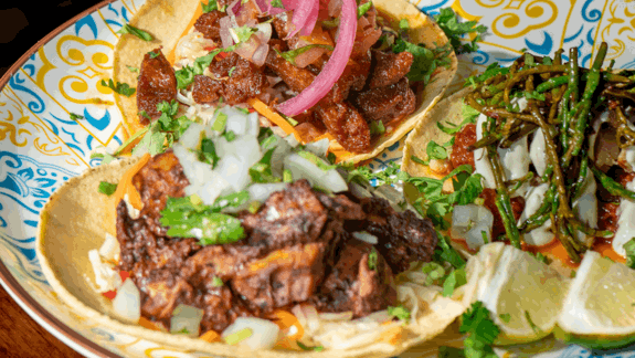 Three vegan Mexican tacos on a colorful plate, topped with grilled samphire, marinated seitan, sautéed vegetables, pickled red onions, cilantro, and lime wedges.