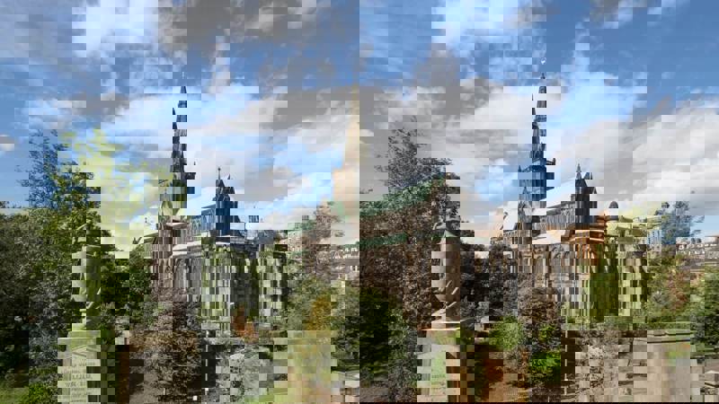 The historic Glasgow Cathedral, a Gothic stone building, stands against a blue sky with white clouds. The foreground includes gravestones and trees.