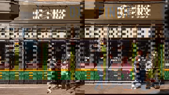 The exterior of a restaurant called "Ox and Finch" with large windows and a green and gold tile facade. People are walking by on the sidewalk.