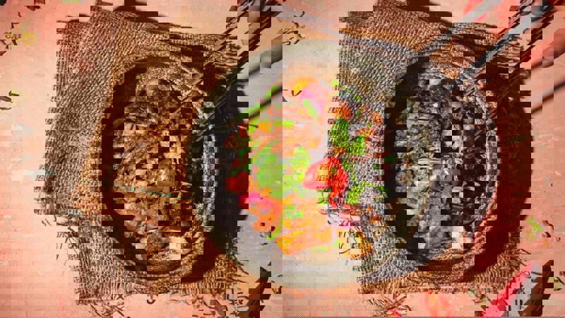 A close-up, overhead shot of a vibrant, spicy stir-fry or curry in a dark bowl, being picked up with chopsticks.