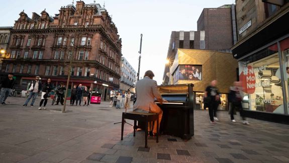 Street performer plays piano on Sauchiehall Street with historic architecture and modern shops. Pedestrians pass by, creating a lively atmosphere.