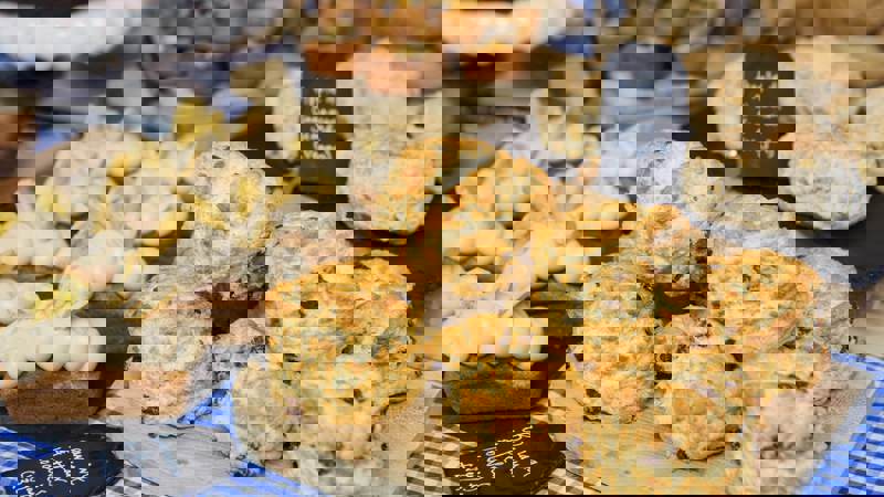 A close-up shows a display of baked goods, including a pile of golden raisin scones on a wooden board, banana and peanut butter loaves, and vegan apple and cinnamon scones, all artfully arranged for sale.