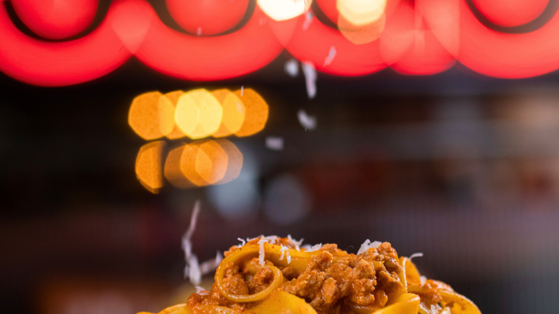 Close-up of fresh parmesan cheese being grated onto a plate of tagliatelle pasta with ragu sauce.