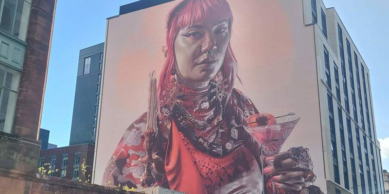 A large, colourful mural on the side of a building, depicting a person with pink hair holding a cocktail and a candle.