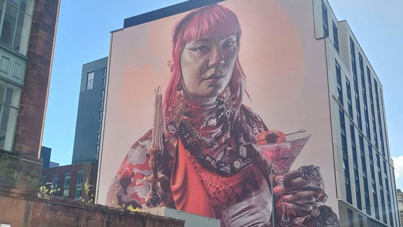 A large, colourful mural on the side of a building, depicting a person with pink hair holding a cocktail and a candle.