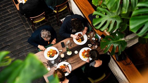 An overhead view of four people dining at a communal wooden table, surrounded by large green plants and cobblestone flooring.